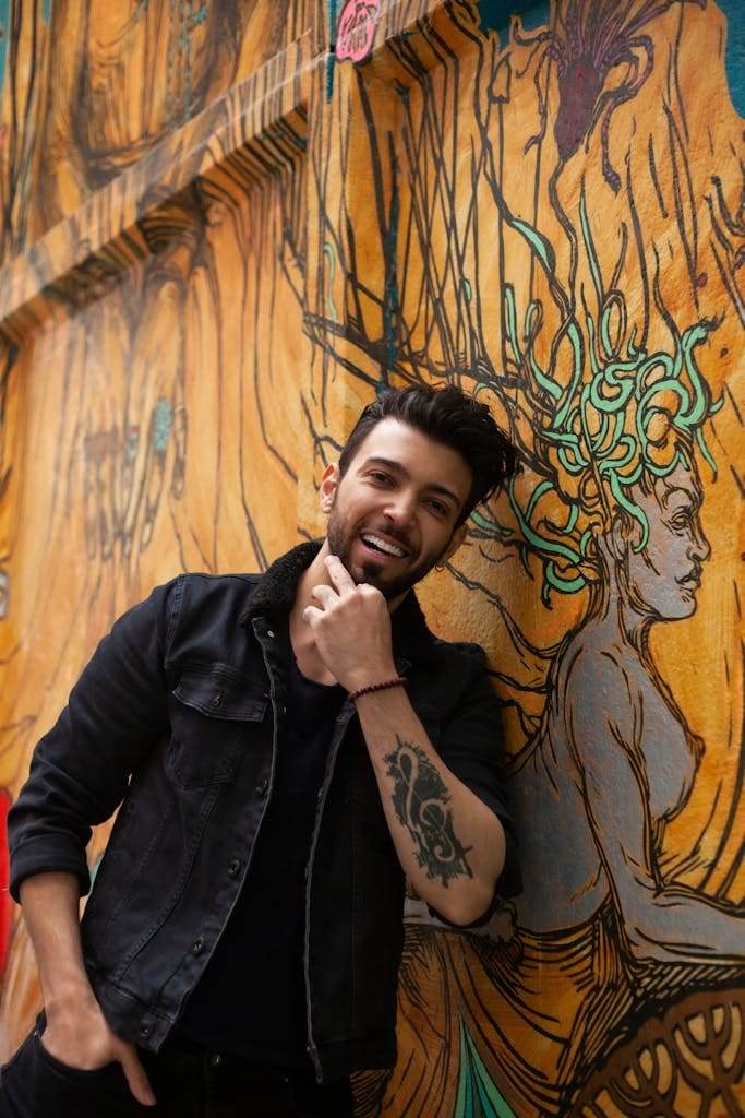 Confident man posing by colorful graffiti wall, showcasing arm tattoo and vibrant expression.